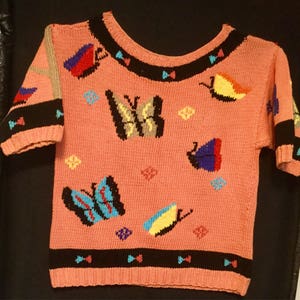 K&ouml;nnte beinhalten: Ein rosa Kurzarm-Pullover mit Rundhalsausschnitt und schwarzem und wei&szlig;em Besatz. Der Pullover ist mit bunten Schmetterlingsmotiven verziert.
