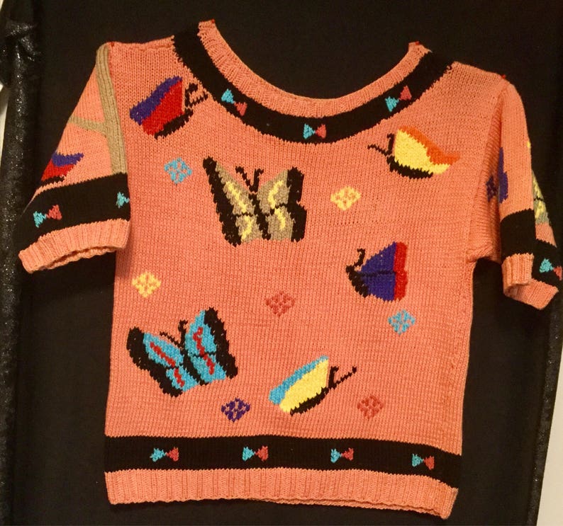 K&ouml;nnte beinhalten: Ein rosa Kurzarm-Pullover mit Rundhalsausschnitt und schwarzem und wei&szlig;em Besatz. Der Pullover ist mit bunten Schmetterlingsmotiven verziert.