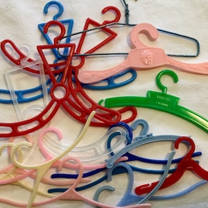 May include: A collection of colorful plastic doll hangers, including pink, red, blue, green, and clear. Some hangers have a decorative design, while others are plain.