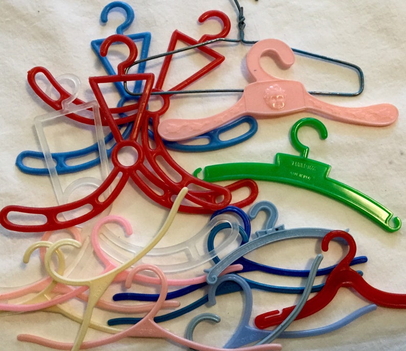 May include: A collection of colorful plastic doll hangers, including pink, red, blue, green, and clear. Some hangers have a decorative design, while others are plain.