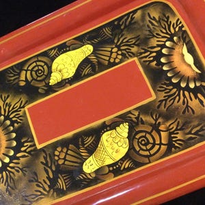 May include: A red wooden tray with a black and gold painted design. The design features seashells and stylized floral patterns.