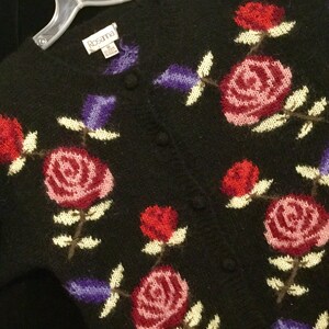 May include: Black knitted cardigan with a floral pattern featuring pink, red, and purple roses. The cardigan has a button closure and the label "Rosanna" is visible on the front.