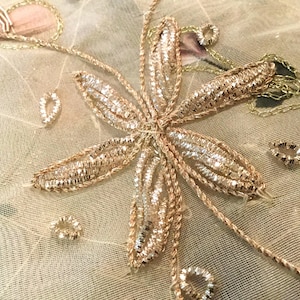 May include: Close-up of a floral embroidery design. The design features a large, six-petal flower with gold and silver thread. Additional small, teardrop-shaped and circular embellishments are scattered around the flower. The background is a light beige fabric.