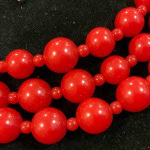 May include: Close-up of a red beaded necklace. The necklace features three strands of large, glossy, round beads connected by smaller beads. The beads are a vibrant, solid red color, and the background is black.
