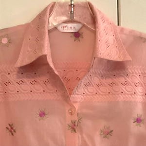 May include: A pale pink button-down shirt with floral embroidery. The shirt has eyelet lace detailing on the collar and yoke. The brand name "PINK" is visible on the label.