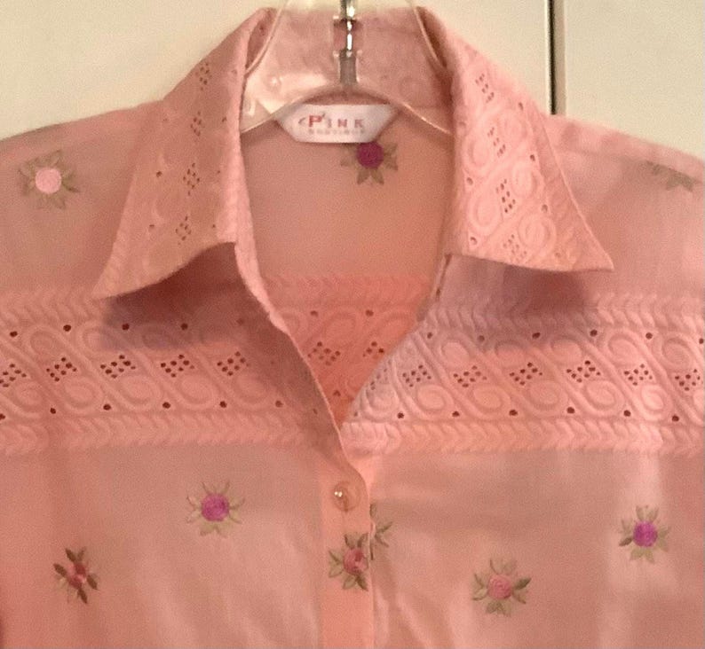 May include: A pale pink button-down shirt with floral embroidery. The shirt has eyelet lace detailing on the collar and yoke. The brand name "PINK" is visible on the label.