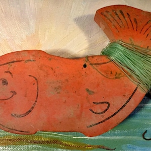 May include: A red wooden fish with a green string wrapped around it. The fish has a black hook attached to it. The fish is painted with black lines to resemble scales and a mouth.