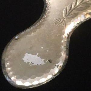 Pu&ograve; includere: Uno specchio da mano argentato con un bordo a festoni e un motivo floreale inciso sulla superficie. Lo specchio ha un aspetto usurato e vintage.