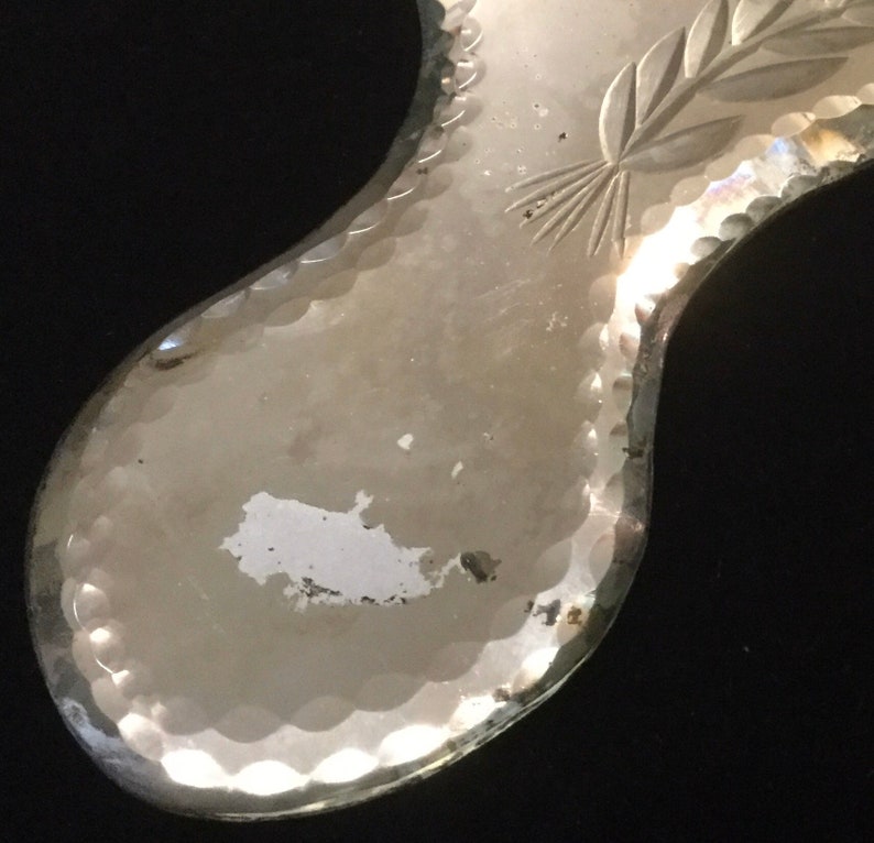 Pu&ograve; includere: Uno specchio da mano argentato con un bordo a festoni e un motivo floreale inciso sulla superficie. Lo specchio ha un aspetto usurato e vintage.