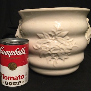 May include: Cream-colored ceramic planter with embossed floral design and two side handles. A can of Campbell's Condensed Tomato Soup is next to the planter. The planter has a wide, rounded shape.