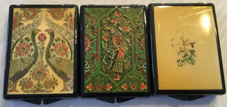 May include: Three decorative compact cases with black frames. The first features two peacocks and floral designs in gold, red, and green. The second has a bird and floral design in green and red. The third has a hummingbird and floral design.