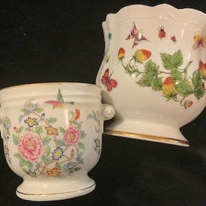 May include: Two white ceramic planters. One features a floral and bird design with pink, blue, and yellow flowers. The other has a strawberry and butterfly motif. Both have gold trim and scalloped edges.