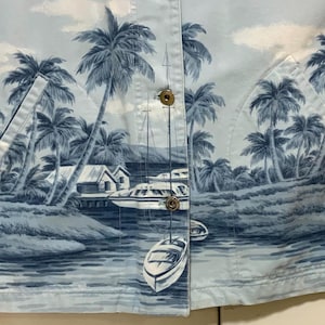 May include: Light blue button-up shirt with a tropical print featuring palm trees, boats, and small buildings. The shirt has two front pockets and gold-colored buttons. The overall design evokes a coastal or vacation theme.