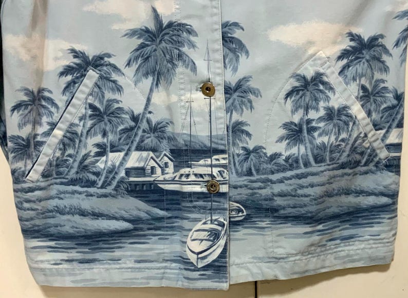 May include: Light blue button-up shirt with a tropical print featuring palm trees, boats, and small buildings. The shirt has two front pockets and gold-colored buttons. The overall design evokes a coastal or vacation theme.