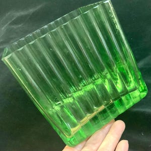 May include: A square, transparent, green glass vase with vertical ribbed detailing. The vase has a rectangular base and is held at an angle, showcasing its textured surface and vibrant color. The glass appears to be vintage.