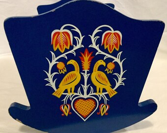 May include: A blue wooden rocking horse with a folk art design featuring two yellow birds, red and white flowers, and a heart. The design is painted on the front of the horse.