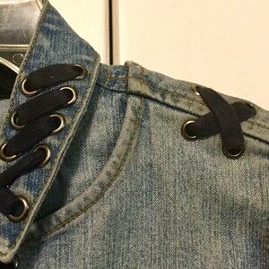 May include: A denim jacket with black lace-up details on the collar and shoulder. The jacket has a distressed look and is made of a soft, comfortable material.