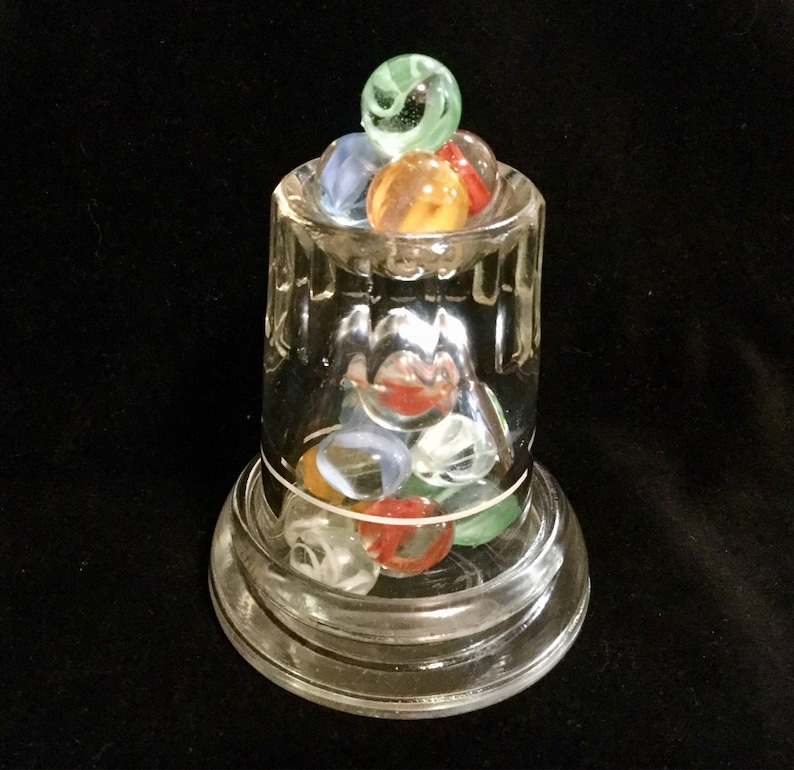 May include: A clear glass jar with a round base and a wide mouth filled with colorful marbles. The marbles are a mix of green, blue, red, and orange.