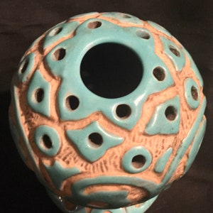 May include: A turquoise ceramic sphere with a large hole in the center and a decorative pattern of white raised lines and holes.