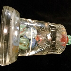 May include: A clear glass bottle with a wide mouth and a textured base. The bottle is filled with colorful marbles, including blue, green, red, and white.