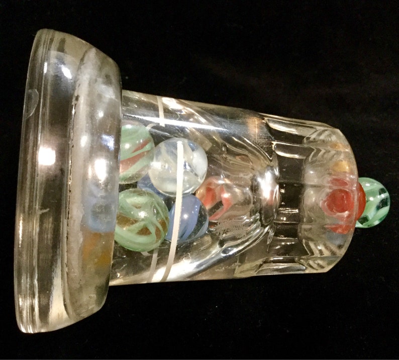 May include: A clear glass bottle with a wide mouth and a textured base. The bottle is filled with colorful marbles, including blue, green, red, and white.