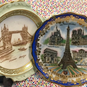 May include: Two decorative ceramic plates with gold trim. The plate on the left features a brown and white illustration of Tower Bridge in London. The plate on the right features a blue and white illustration of the Eiffel Tower in Paris with the text "La Tour Eiffel", "Le Sacré-Coeur", "Notre-Dame", "Arc de Triomphe", "Le Louvre", and "SOUVENIR de PARIS".