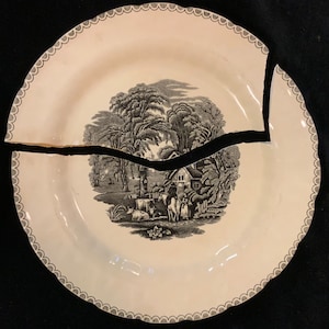 May include: A broken white ceramic plate with a black and white pastoral scene. The plate is in two pieces, with a large crack running across the center. The rim has a scalloped edge with a gray border. The scene depicts a house, trees, and figures.