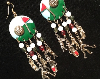 May include: Gold tone golf-themed dangle earrings with green, white, and red enamel. The earrings feature a golf ball, flag, and green, and dangle with charms of golfers, golf carts, and golf clubs.