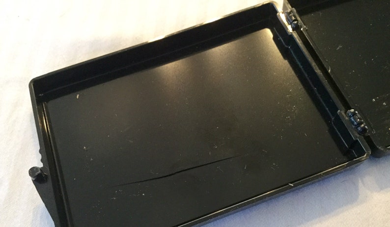 May include: An open, black, rectangular case with a hinged lid. The interior of the case is black and has a crack running across the bottom. The case is on a white surface.