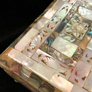 May include: Close-up of a decorative box with a mosaic design. The box is covered in iridescent mother-of-pearl tiles, with carved floral and leaf accents. The tiles are arranged in a geometric pattern, creating a shimmering effect.