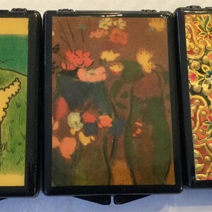May include: Three small, black hinged boxes with decorative tops. One features a yellow sheep in a green field, another a floral still life, and the third an ornate gold and red pattern. The boxes are likely for small items.