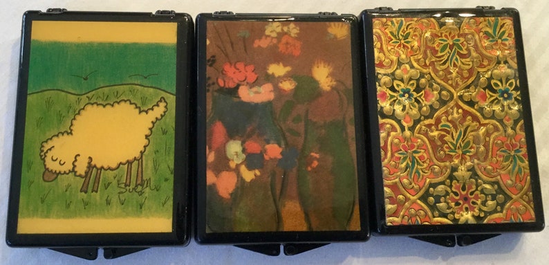 May include: Three small, black hinged boxes with decorative tops. One features a yellow sheep in a green field, another a floral still life, and the third an ornate gold and red pattern. The boxes are likely for small items.