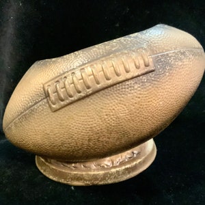 May include: A bronze-colored football-shaped decorative piece. The football has a textured surface and a raised panel with vertical lines. It sits on a round base with a decorative edge. The piece is likely a sports-themed home decor item.