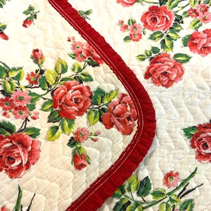 May include: A quilted textile featuring a floral pattern of red roses and green leaves on a cream background. A red, ruffled trim borders the edge. The fabric has a diamond-shaped quilted pattern.