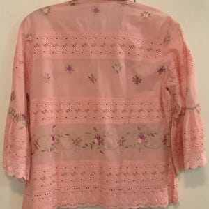 May include: A pale pink blouse with long sleeves, detailed with eyelet and floral embroidery. The blouse has a collared neckline and flared sleeves. The design incorporates floral embroidery and scalloped edges, offering a delicate, feminine style.