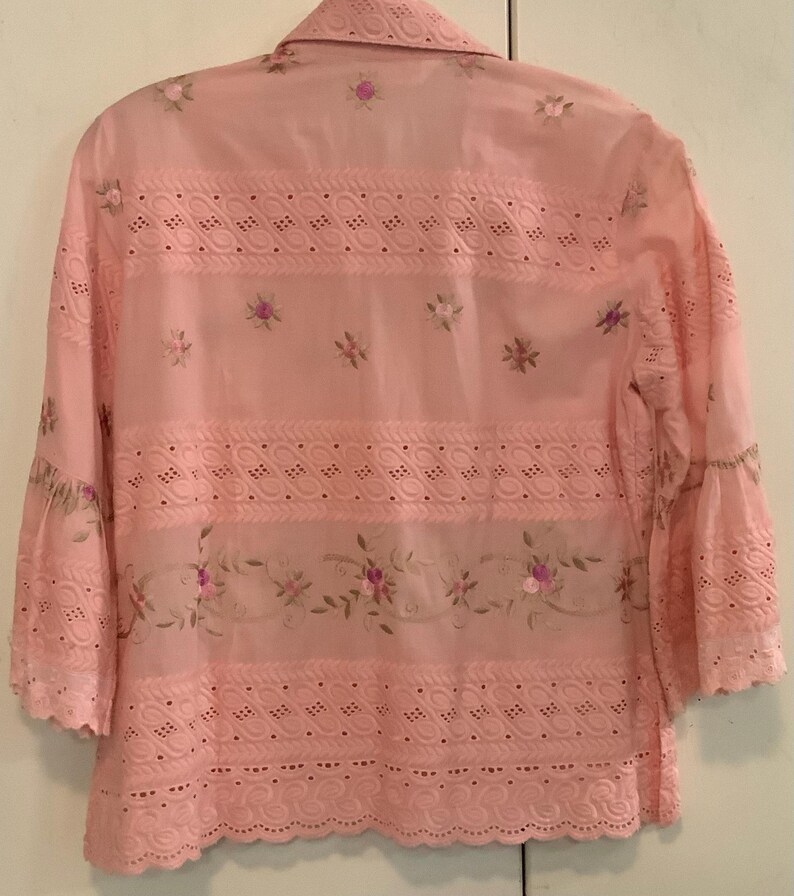 May include: A pale pink blouse with long sleeves, detailed with eyelet and floral embroidery. The blouse has a collared neckline and flared sleeves. The design incorporates floral embroidery and scalloped edges, offering a delicate, feminine style.
