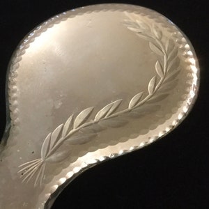 Pu&ograve; includere: Uno specchio da mano in argento con un design inciso decorativo di una ghirlanda di foglie attorno al bordo.