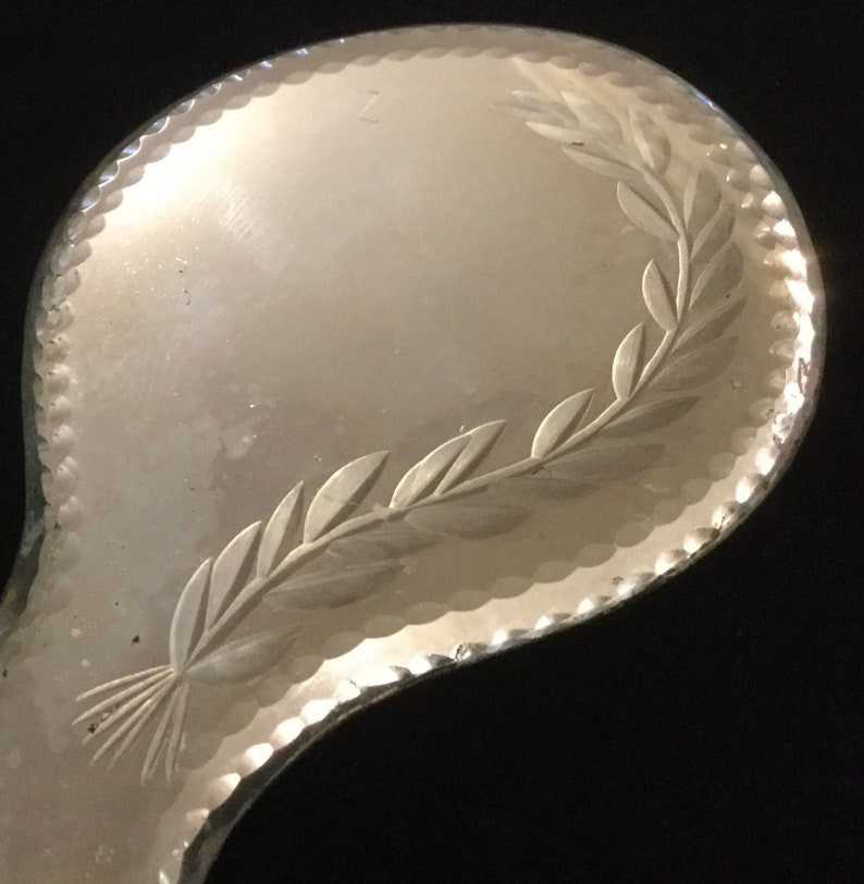 Pu&ograve; includere: Uno specchio da mano in argento con un design inciso decorativo di una ghirlanda di foglie attorno al bordo.