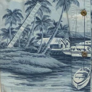 May include: Light blue button-up shirt with a tropical scene print. The print features palm trees, small houses, boats, and water in shades of blue and white. The shirt has a pocket and gold-colored buttons.