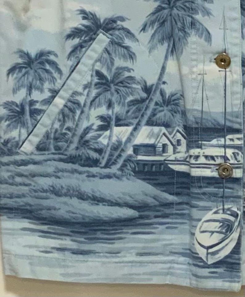 May include: Light blue button-up shirt with a tropical scene print. The print features palm trees, small houses, boats, and water in shades of blue and white. The shirt has a pocket and gold-colored buttons.