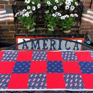 May include: A red, white, and blue patchwork table runner with a patriotic theme. The runner features alternating red squares and blue squares with white anchor and flag patterns. The word "AMERICA" is visible in the background.