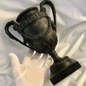 May include: A dark bronze, ornate, two-handled vase with a square base. The vase is decorated with intricate floral and leaf designs.