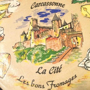 May include: A round ceramic plate with a colorful illustration of a medieval castle, the text "Carcassonne La Cité Les bons Fromages", and various types of cheese.