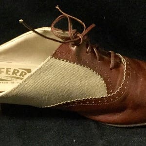 May include: Brown leather slip-on shoe with a beige canvas panel and white stitching. The shoe has a lace-up design and the brand name "FERR" is visible on the insole.