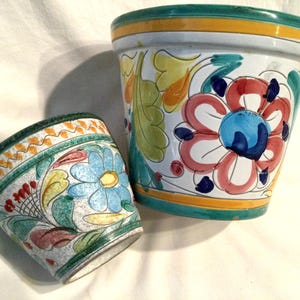 May include: Two ceramic flower pots with colorful floral designs. The larger pot is white with green, yellow, blue, and red flowers. The smaller pot is white with green, yellow, blue, and pink flowers.
