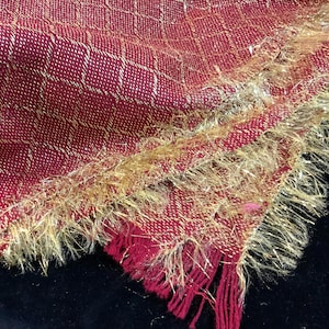 May include: Close-up of a burgundy scarf with a woven grid pattern and gold metallic threads. The scarf has a fringed edge with gold and burgundy threads, creating a textured and elegant look. The scarf is made of a soft material.