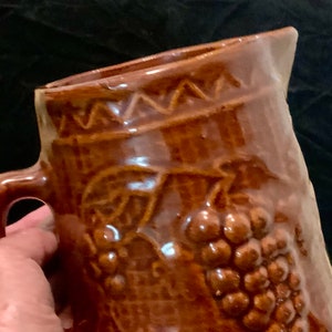 May include: Brown ceramic pitcher with a grape design. The pitcher has a handle and a textured surface.