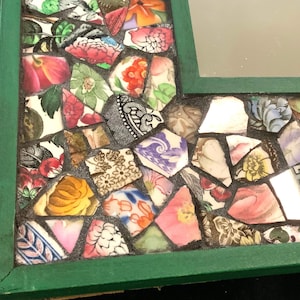 May include: A green framed mirror with a mosaic border made of broken pieces of colorful china. The mosaic features floral patterns and various colors, including pink, blue, yellow, and green.