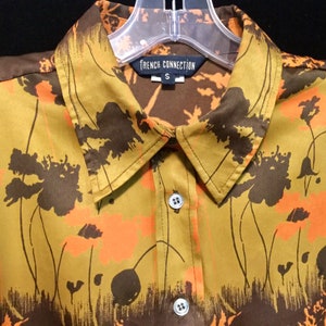 May include: A mustard yellow button-down shirt with a floral print in shades of brown and orange. The shirt has a collar and a button closure. The label reads "French Connection" and "S".