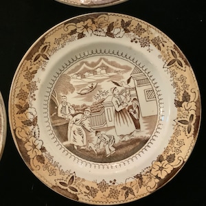 May include: A white ceramic plate with a brown floral border and a brown and white scene of people in a landscape. The scene includes a house, a boat, and trees.